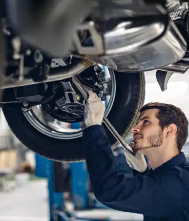 A mechanic wearing gloves works underneath a raised vehicle, using a wrench to adjust parts near the car’s rear wheel.