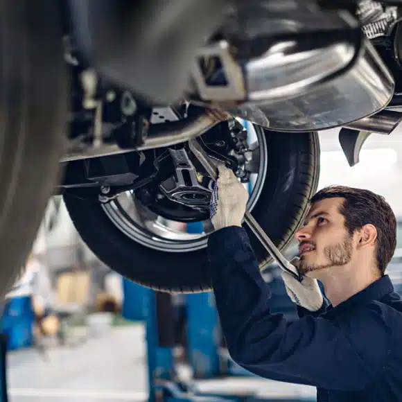 A mechanic wearing gloves and a blue uniform uses a wrench to work on the underside of a car elevated on a lift.